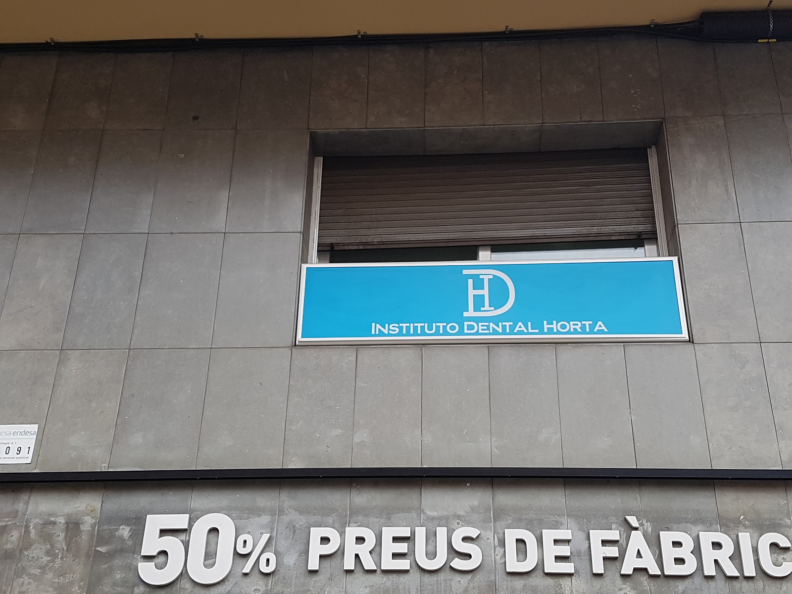 Fachada de losas de hormigón gris. Sobre una ventana con persiana, un rótulo azul celeste con el monograma HD y el texto INSTITUTO DENTAL HORTA; más abajo, pasada una franja negra, letras blancas condensadas montadas sobre el hormigón que dicen 50% PREUS DE FÀBRICA, con la última letra fuera del encuadre.