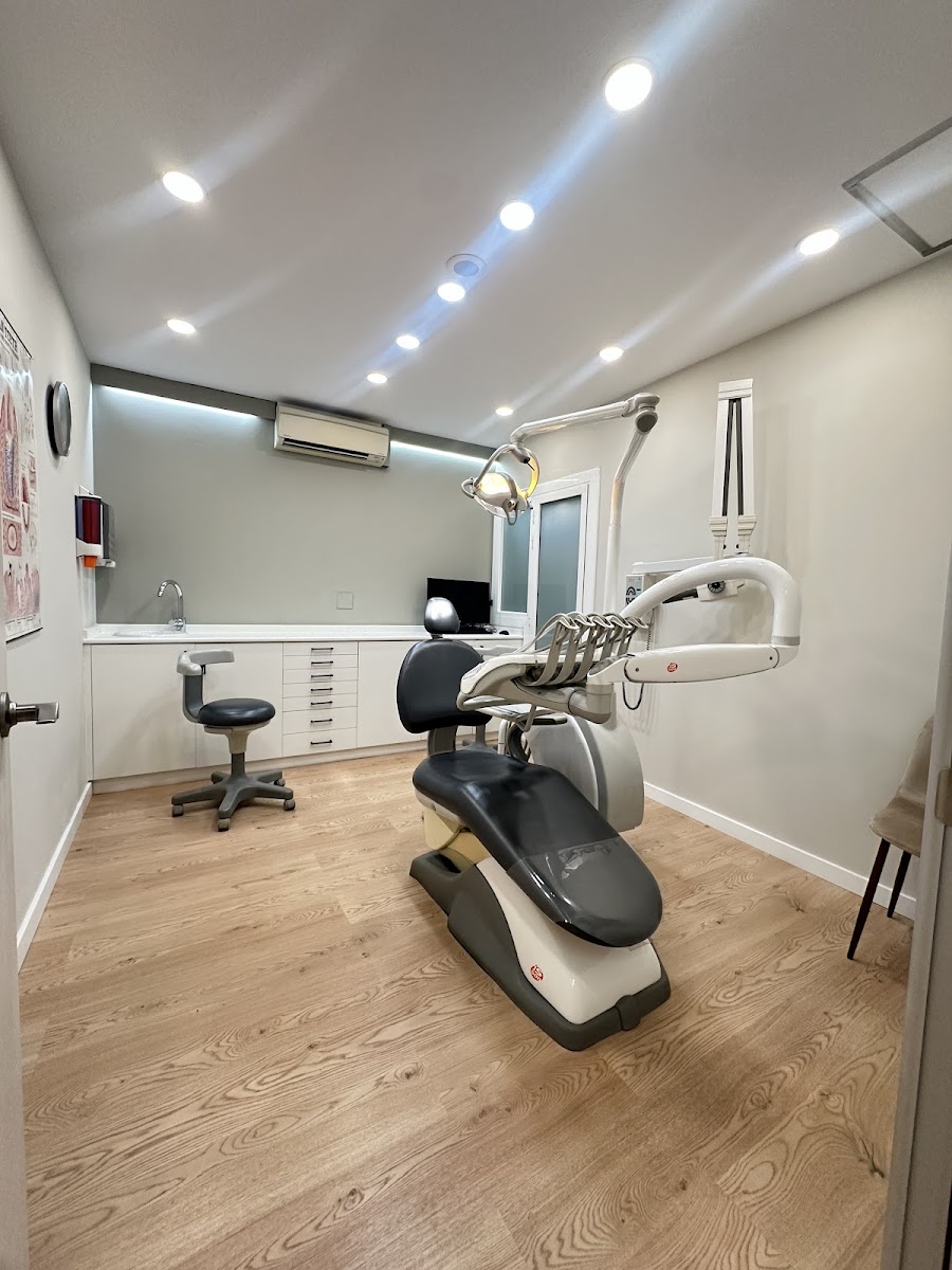 Sala de tratamiento con suelo de madera de roble claro: sillón dental gris y negro con su lámpara, taburete, encimera blanca con pica y cajonera, aire acondicionado en la pared y focos empotrados en el techo.