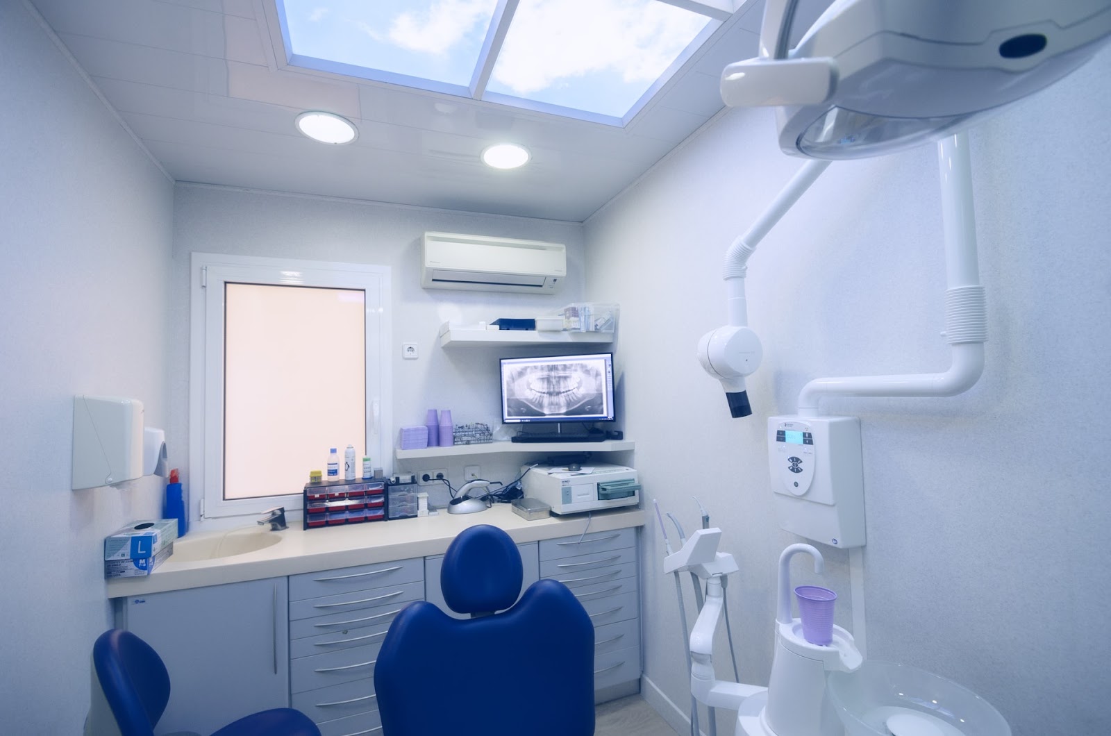 Sala de tratamiento de paredes blancas: sillón tapizado en azul, mobiliario gris claro con encimera y pica, un monitor con una radiografía panorámica, un equipo de rayos montado en la pared y un panel de techo retroiluminado con un cielo con nubes.